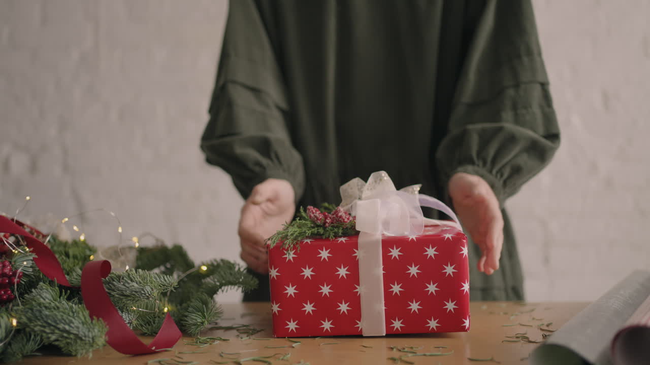 A Christmas present moves a woman's hands into the camera. Red Packaged Gift Woman Moves Into Camera After Packing and Decorating