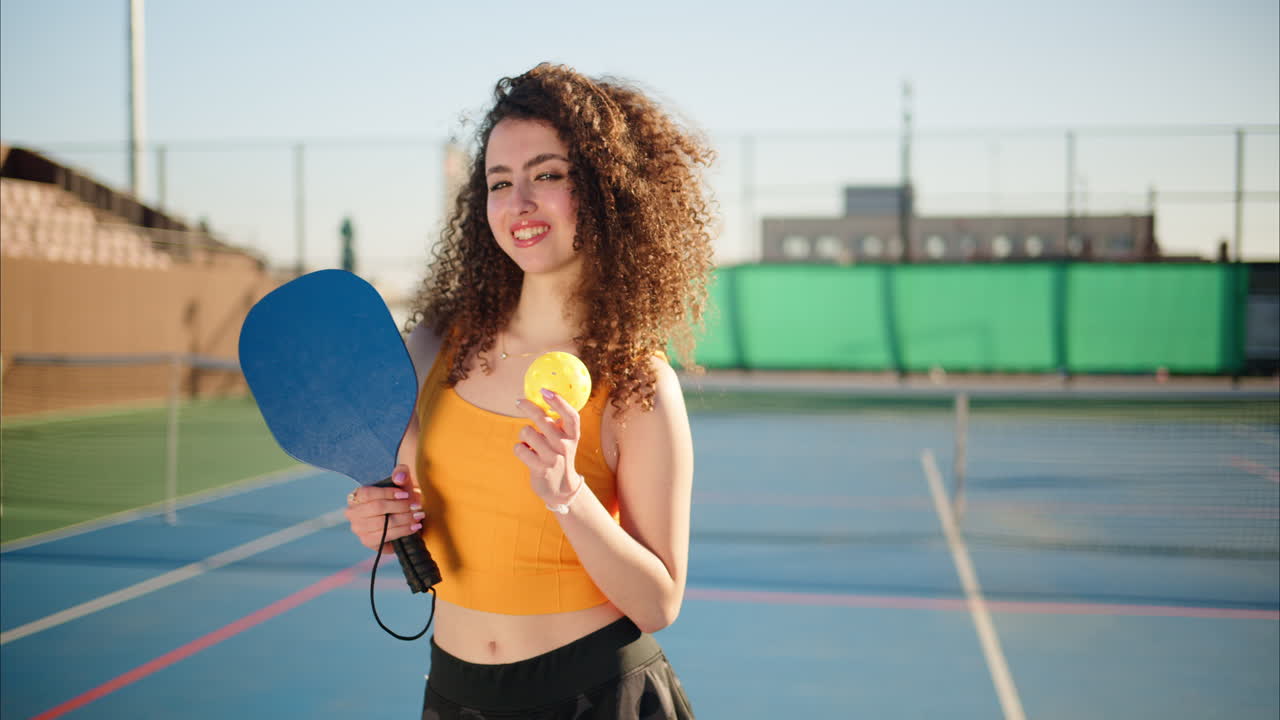A happy and smiling woman with curly hair holding a Pickleball blue racket and a yellow ball on a blue court on a sunny day