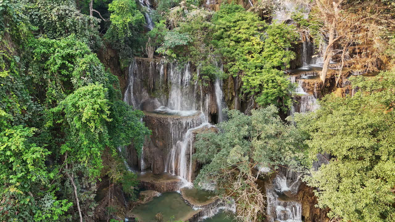 Vista aerea con drone della cascata principale di Kuang Si vicino a Luang Prabang, dove molteplici corsi d'acqua precipitano da formazioni rocciose calcaree in pozze turchesi circondate da una giungla fitta.