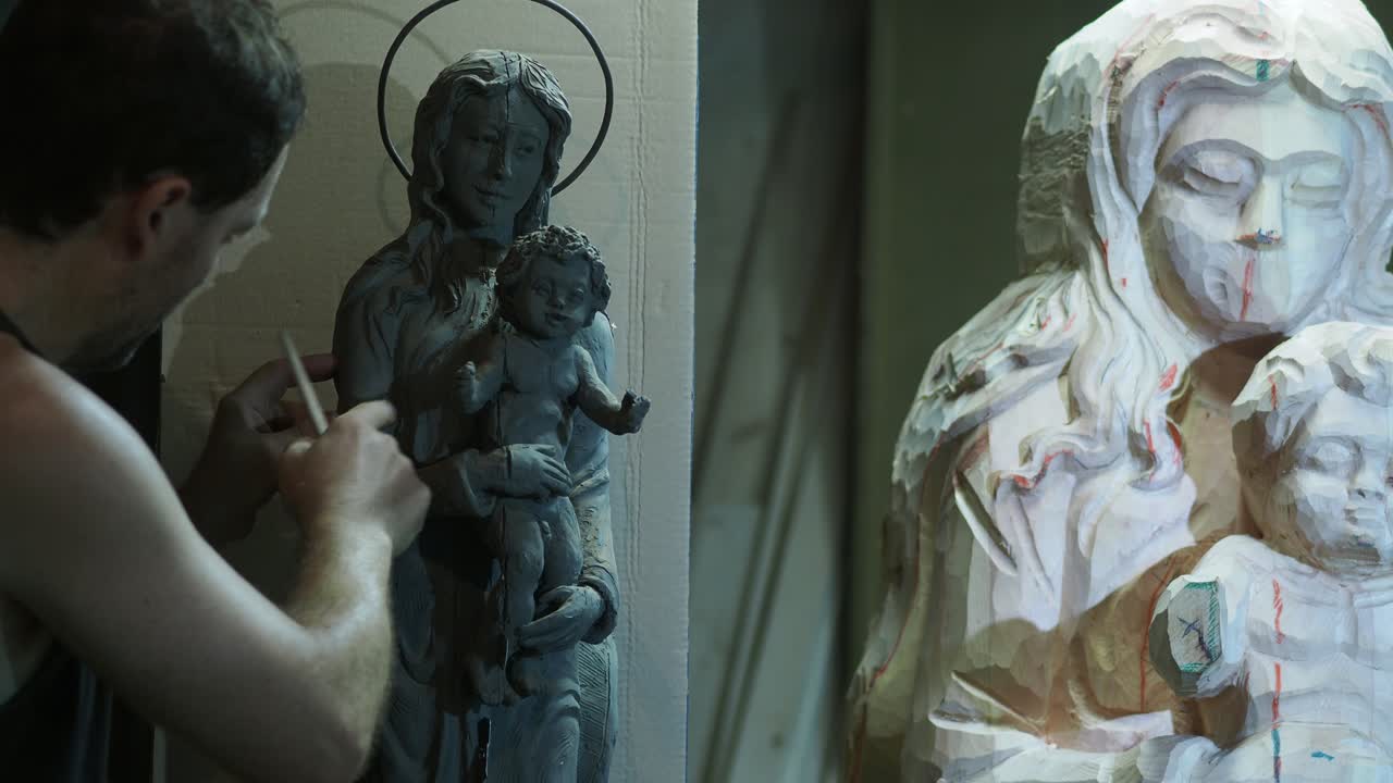Artist Meticulously refining scaled clay model near wooden Religious Madonna and Child Statue work in progress in Workshop Detailed Craftsmanship