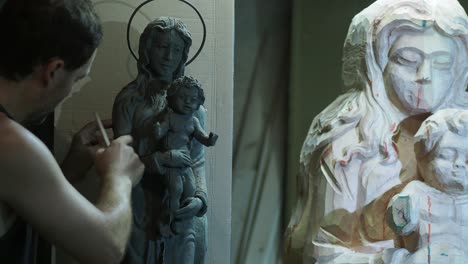 artist meticulously refining scaled clay model near wooden religious madonna and child statue work in progress in workshop detailed craftsmanship