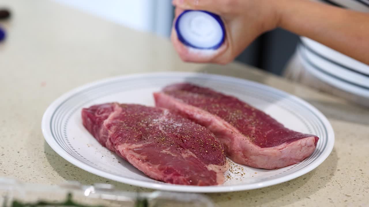 Close-up of hands seasoning beef steaks with salt and pepper in a kitchen setting