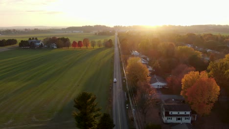 car drives on road that bisects rural farmland and neighborhood community homes during colorful sunrise, sunset