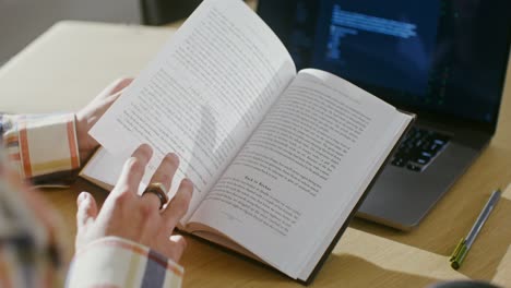 Person reading a book while looking at a laptop with code on the screen