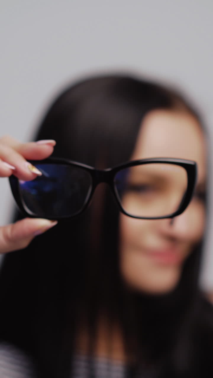 Pretty woman shows from left to right stylish eyeglasses and puts them on with blur background. Fashionable glasses in the hand of dark-haired female. Close-up Vertical video