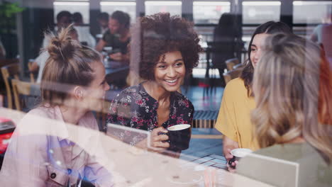 Four-Young-Female-Friends-Meeting-Sitting-At-Table-In-Coffee-Shop-And-Talking-Viewed-Through-Window