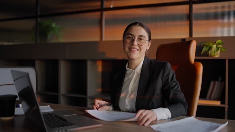 portrait of a happy brunette businesswoman in round glasses and business uniform sitting at a table in front of a laptop and working in a sunny office