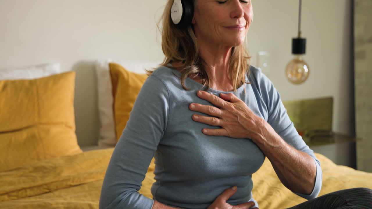 Senior woman meditating with headphones, relaxing on bed at home