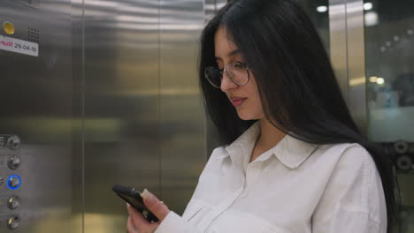 marketer wearing white shirt chats on phone with client while riding elevator in modern building interior, smiling warmly and holding railing, surrounded by reflective metal walls