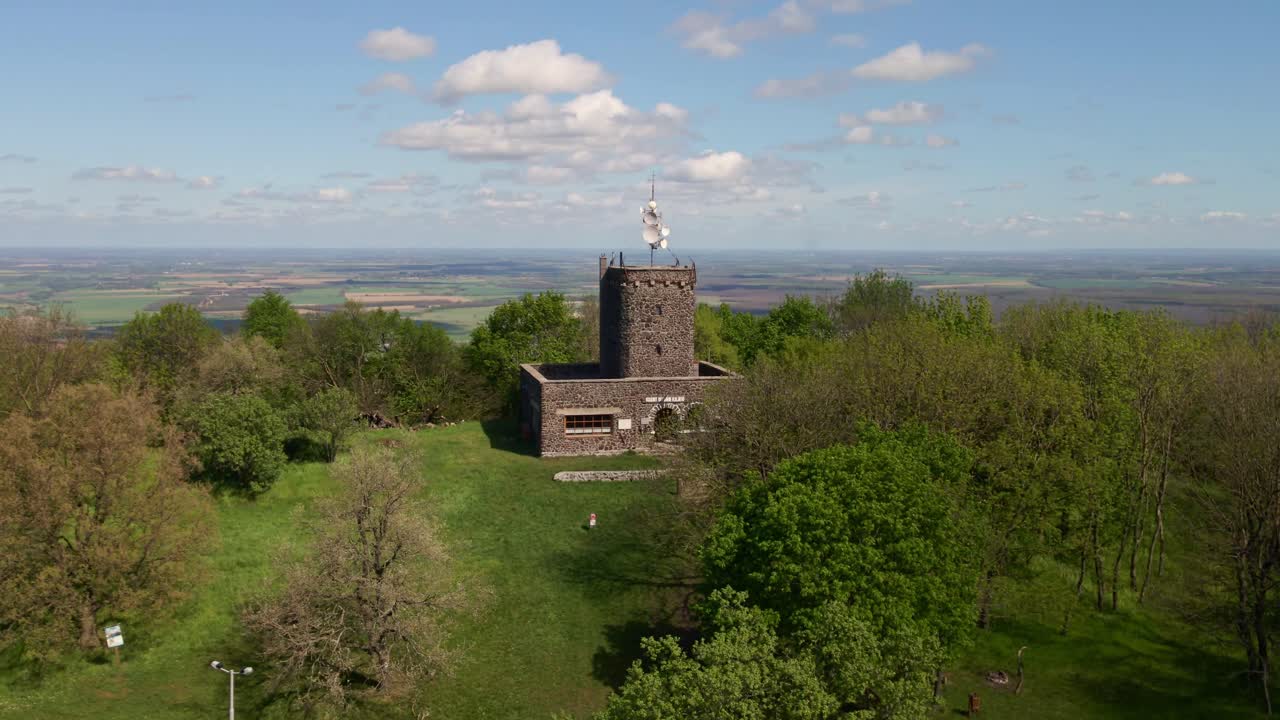 Aerial view of a historic stone observation tower nestled within a vibrant green forest, set against a backdrop of vast open fields and clear blue skies.
