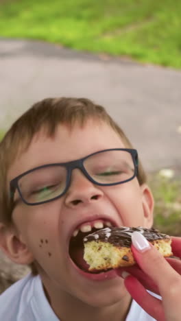 un momento lúdico entre una madre y su hijo, donde ella le ofrece un pastel de chocolate mientras está sentado en un campo cubierto de hierba bajo un árbol, el niño felizmente abre la boca para un bocado