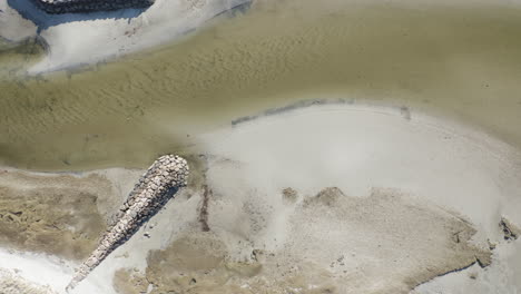 the camera looks down and gives a birds eye view of rock harbor (orleans, ma) marina at low tide. there is a small exposed shoreline due to the low tide. docked boats can be seen along the marina path