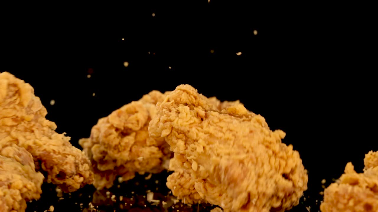 Golden fried chicken pieces fall and bounce on a glossy black background, captured in slow motion with dramatic lighting and visible crumbs scattering