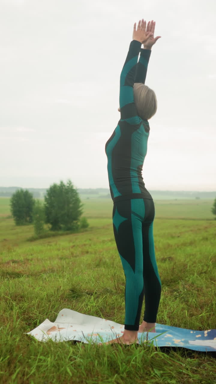 mujer de mediana edad de pie en una alfombra de yoga con los brazos extendidos levantados hacia el cielo, preparándose para una postura de flexión hacia adelante en un campo de hierba brumosa, bajo un cielo nublado con árboles alineados en la distancia