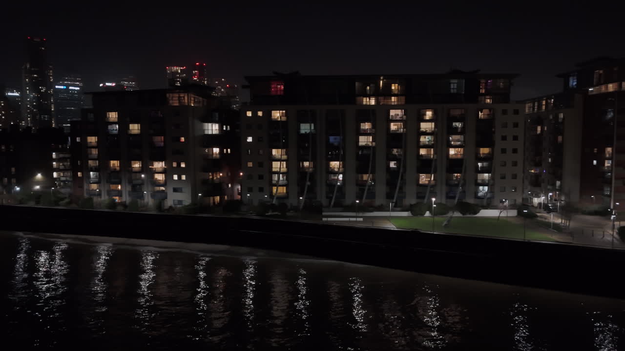 Aerial tracking shot of illuminated riverfront apartments, in Isle of Dogs, London