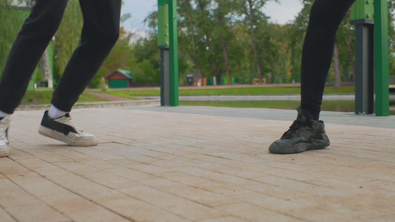 park pavement closeup dancing feet perform rapid shuffle duel on tiled promenade, two young dancers in contrasting sneakers execute precise tapping and footwork, willow trees and green posts frame
