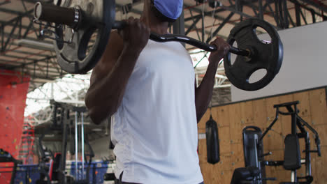 Fit-african-american-man-wearing-face-mask-exercising-using-barbell-in-the-gym