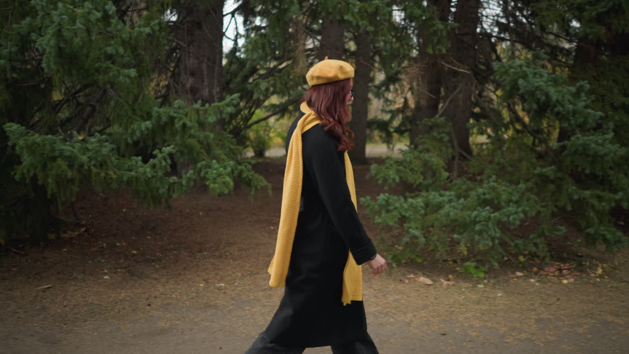 estudiante emocionada con largo cabello rojo ondulado con un abrigo negro, boina amarilla, y bufanda a juego, caminando a través de un exuberante parque verde con colorido follaje de otoño, felizmente levantando la mano a la cabeza