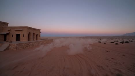 sand swirling around a desert house at sunset creates a stunning and tranquil atmosphere. golden hues fill the sky, enhancing the serene beauty of this remote landscape