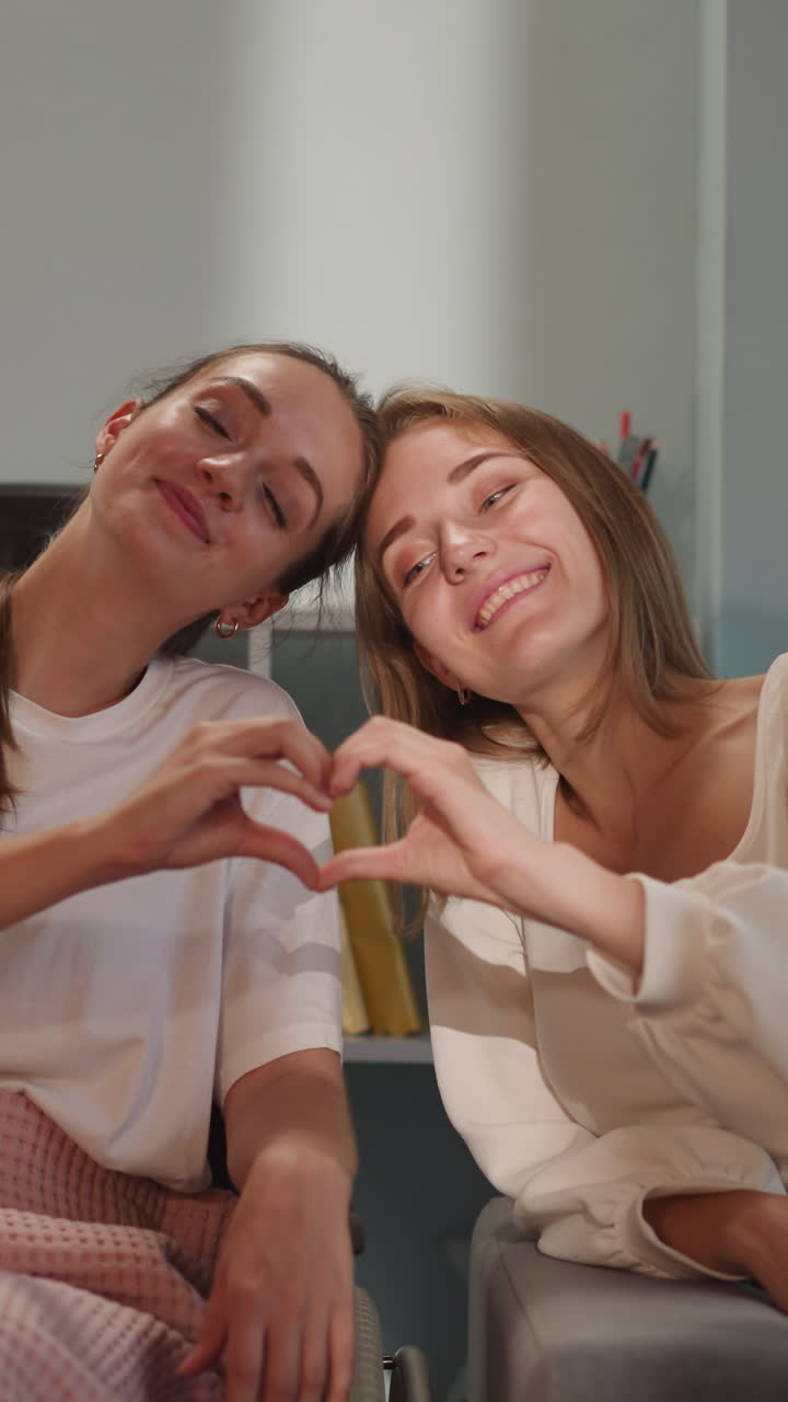 las lesbianas hacen forma de corazón con los dedos sonriendo y mirando a la cámara. mujer feliz con discapacidad en silla de ruedas y novia rubia muestran amor