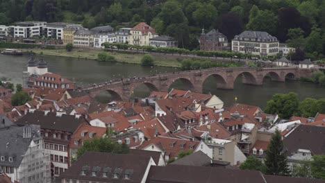 Heidelberg-old-town-rooftops-and-Karl-Theodor-arch-bridge-over-Neckar-River,-hillside-covered-in-dense-woods-in-background,-Germany