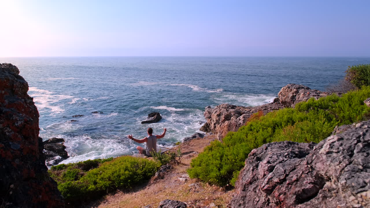 Seated man on sea cliff does morning meditation breathing and wellness flow