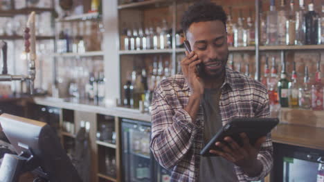 Male-Bar-Worker-On-Phone-Standing-Behind-Counter-Ordering-Stock-Using-Digital-Tablet