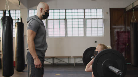 Fitte,-Kaukasische-Frau-Mit-Gesichtsmaske,-Die-Mit-Einer-Langhantel-Mit-Einem-Männlichen-Trainer-Im-Fitnessstudio-Trainiert