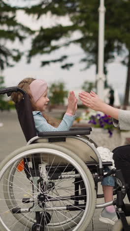 mother and daughter with spinal paralysis play patty in city park. young woman sits on bench entertaining little girl in wheelchair on spring day