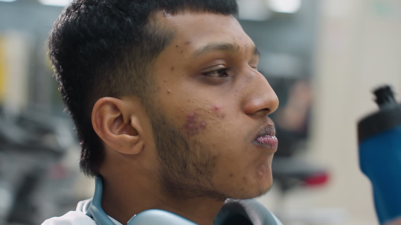 young man wearing white long sleeve top and headphones around neck lifting blue bottle to drink juice in gym environment with blurred workout machines and soft lighting in background