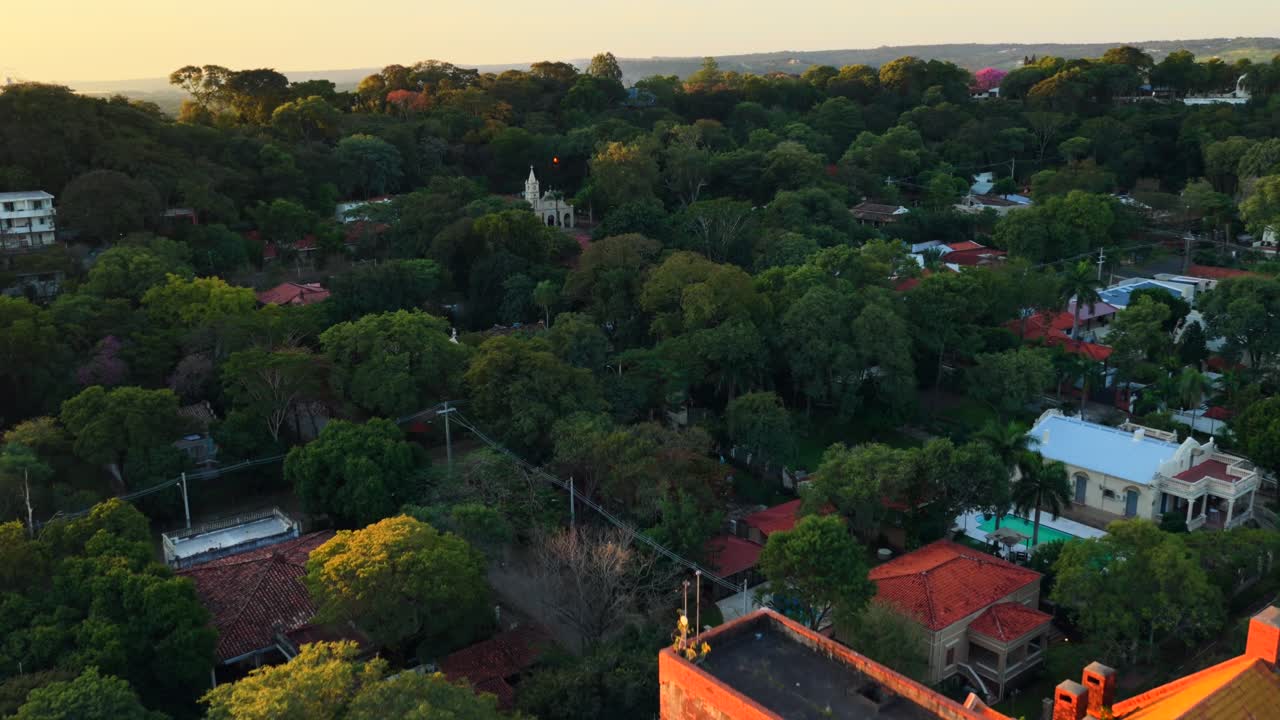 Vídeo com drone da arquitetura marcante de tijolos vermelhos banhada pelo brilho dourado do pôr do sol em San Bernardino, Paraguai. A vegetação exuberante e a atmosfera serena realçam a vista panorâmica.
