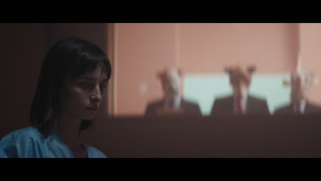 a girl sits at a table and strange figures in animal masks observes her from behind the glass