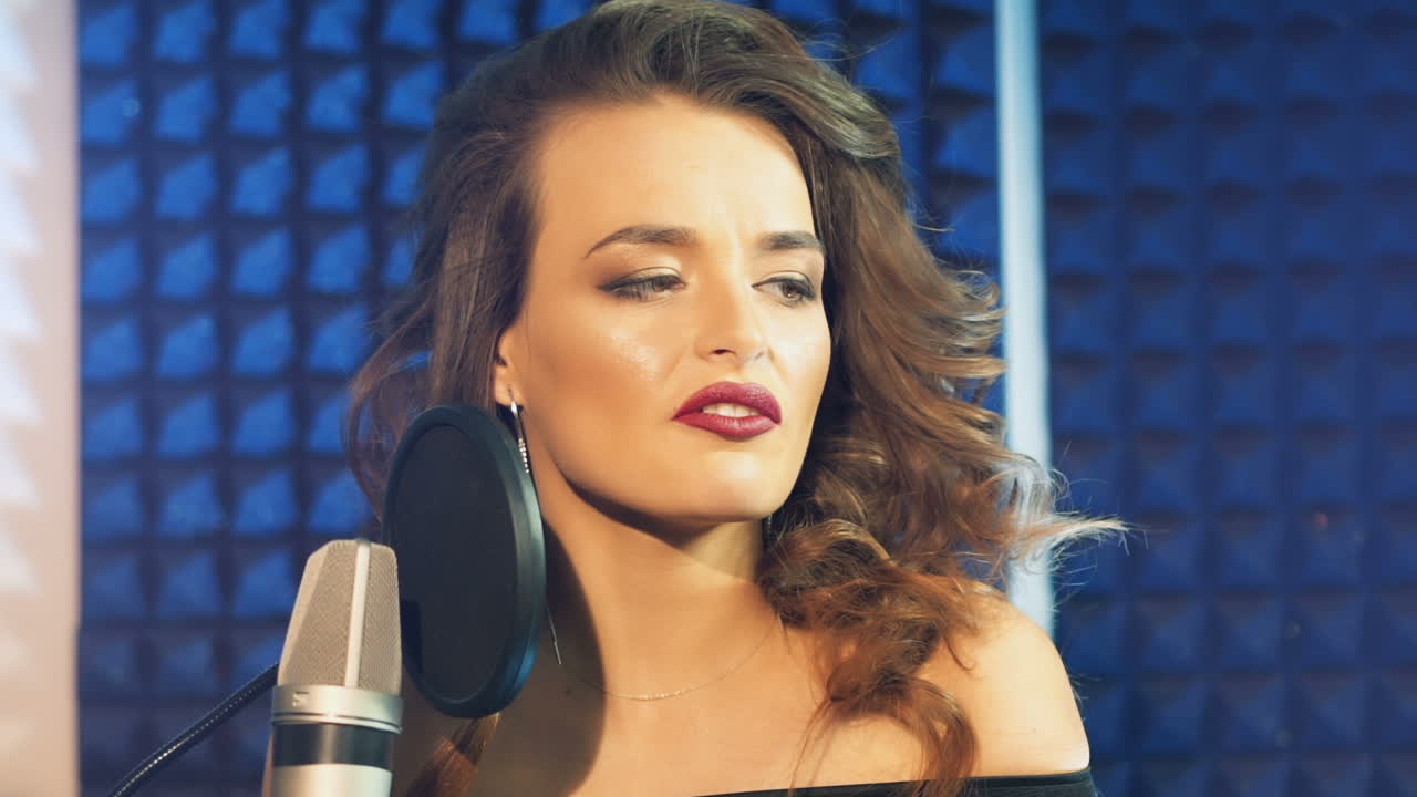 Attractive singer with dark wavy hair and bare shoulders singing into the mic over the blue background. Beautiful woman in a recording studio. Close-up