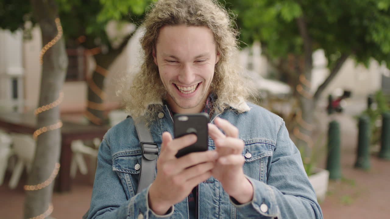 ritratto di un attraente giovane studente maschio biondo che scrive messaggi navigando utilizzando uno smartphone sorridendo felice godendo della tecnologia mobile in una città urbana