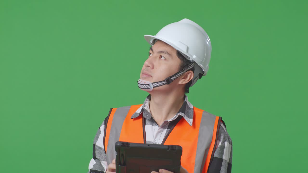 Close Up Of Asian Male Engineer With Safety Helmet Looking At The Tablet In His Hand And Looking Around While Standing In The Green Screen Background Studio
