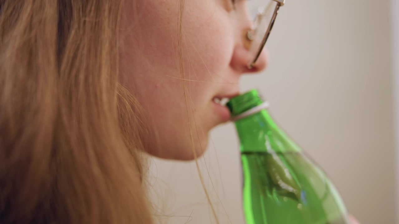 Mulher branca sorrindo enquanto bebe de uma garrafa verde, momento de hidratação alegre em casa aconchegante, óculos brilhando, gole suave e expressão satisfeita, vibração de estilo de vida relaxado, close-up na boca e na garrafa
