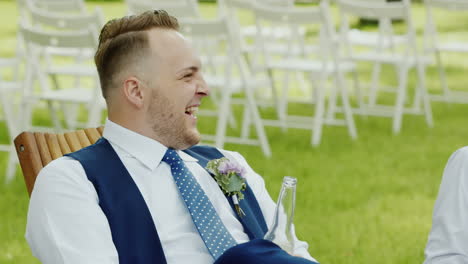 A-Stylish-Man-In-A-Suit-Sits-On-A-Wooden-Chair-In-The-Street-Smiling-Holding-A-Bottle-Of-Beer-On-The