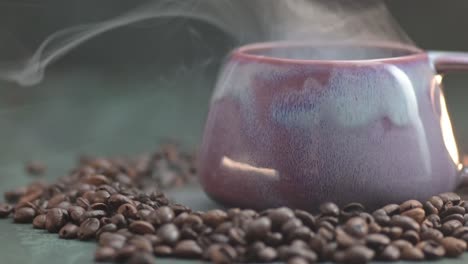 Zoom-in-macro-of-a-smoke-drifts-over-the-lilac-cup,-mug-surrounded-by-laying-on-the-table-coffee-beans