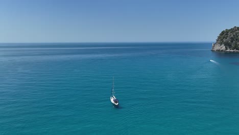 Sailboat-cruising-the-turquoise-waters-near-the-rocky-coast-of-Corfu-Island,-Greece,-under-a-clear-blue-sky