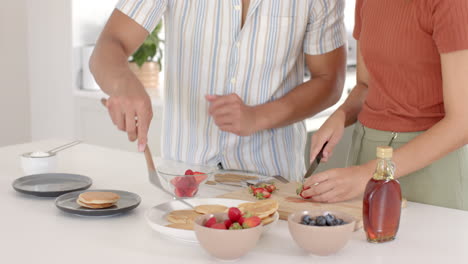 Diverse couple cooking together in kitchen, slicing fruit and flipping pancakes happily, at home