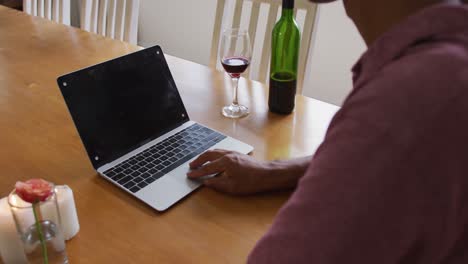 sección media de un hombre de raza mixta sentado en la mesa haciendo una llamada de video con una computadora portátil y saludando