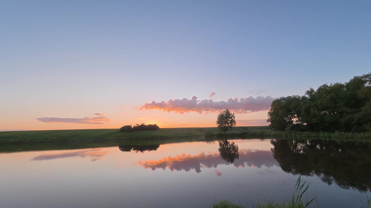 A time lapse captures a beautiful sunset over a calm lake, reflecting the vibrant sky and a lone tree silhouette on the horizon