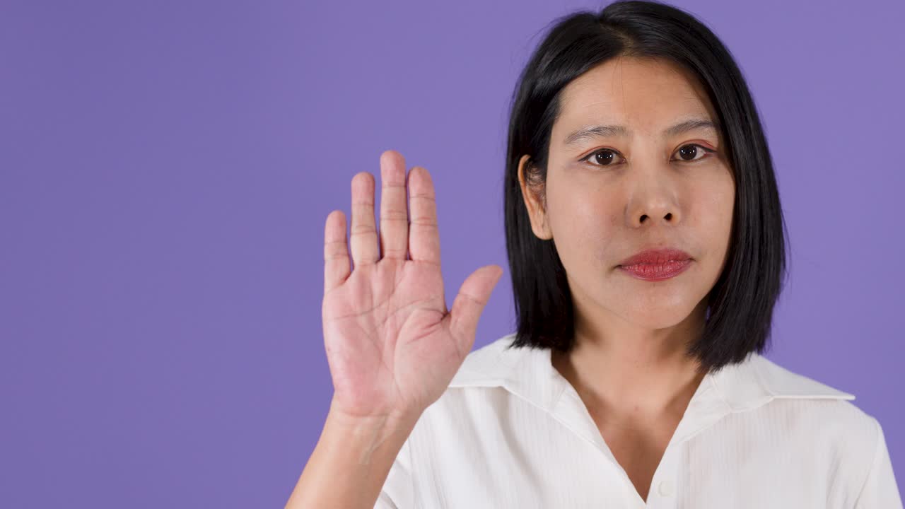 A serious Asian woman raises her hand in a stop gesture against purple studio background