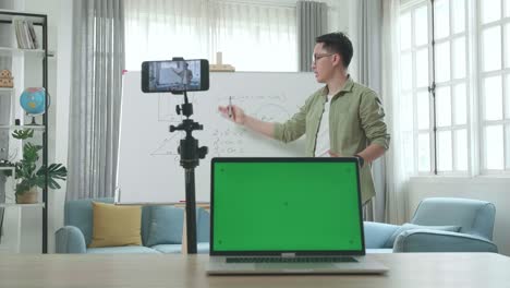 asian male teacher with glasses and green screen laptop shoots video by smartphone camera while teaching math at home