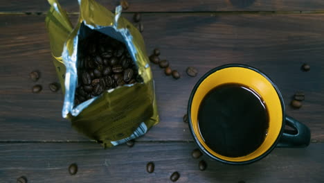 top shot of a cup of steaming coffee next to a packet of coffee beans and coffe beans on a wooden table