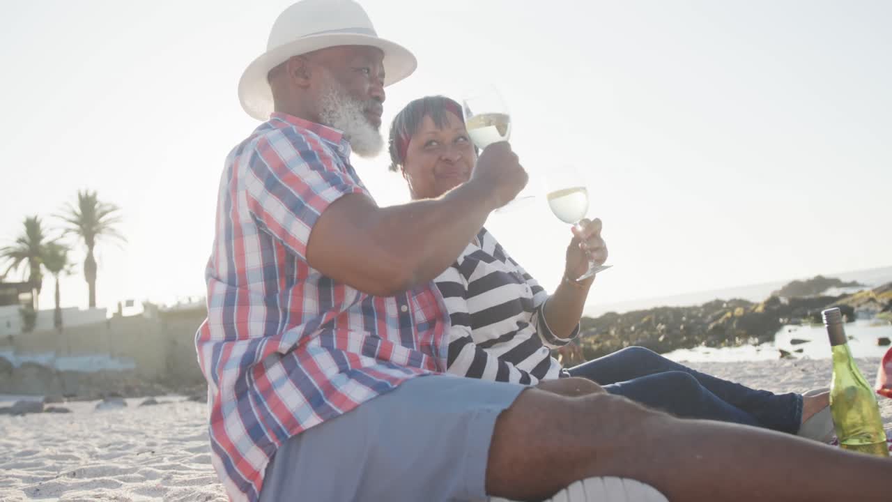 Happy senior african american couple having picnic at beach, drinking white wine, slow motion