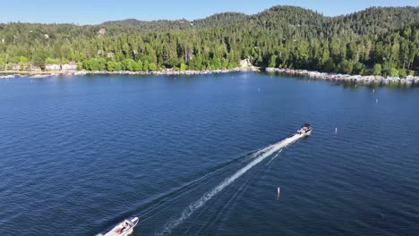 speedboat-and-pontoon-on-lake-arrowhead-in-the-middle-of-california-summer-time-bright-blue-water-and-lakeside-homes-visible-AERIAL-TRUCKING-PAN-FOLLOW