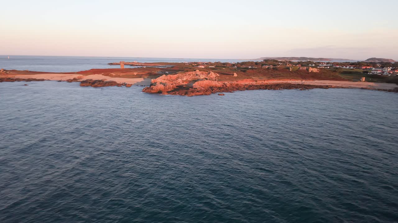 Flight pulling back across the calm waters of Lancresse Bay Guernsey with calm sea showing shoreline and headland basking in golden hour sunshine