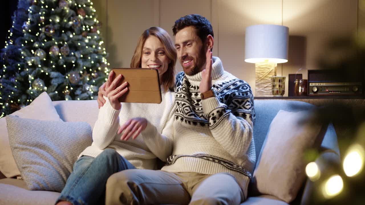 una alegre pareja familiar sonriente saludando con la mano y charlando en videollamadas en línea en una tableta con amigos mientras se sienta en la habitación con un árbol de navidad en la víspera de año nuevo por la noche concepto de espíritu navideño