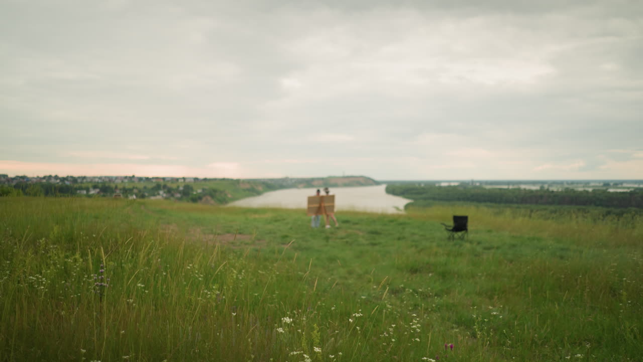 en un campo verde de hierba alta con flores silvestres, el fondo está intencionalmente borroso, revelando una escena serena a orillas del lago. dos figuras distantes y una silla solitaria cerca del lago